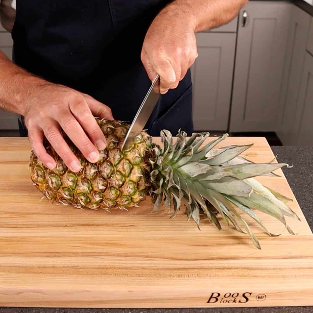 Pineapple on its side laying on a cutting board with a knife cutting off the leaves.